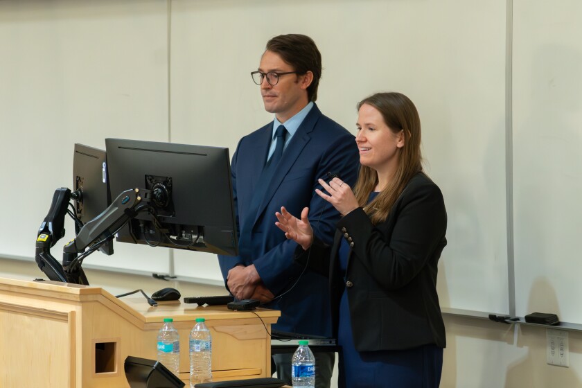 Dr. Jaramillo stands to the left. He is wearing a crisp navy suit. Dr. Orgill stands to the left, she is speaking animatedly and gesturing with one hand while she holds a small, handheld microphone in the other.