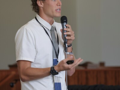 A young man wearing a white shirt and blue tie speaks into a microphone while holding a small device in his other hand.
