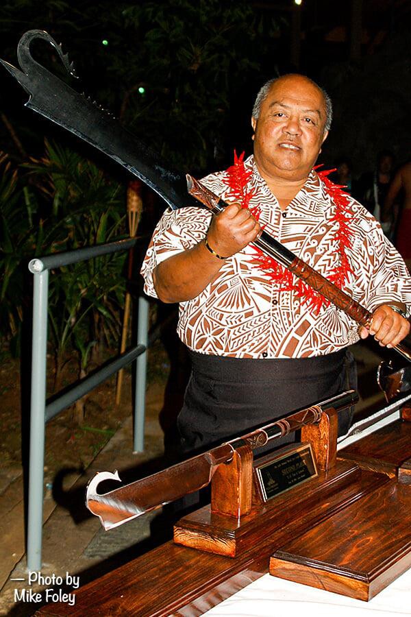 Pulefano Galea'i dressed in a Hawaiian shirt and red lei holds a Samoan fire knife next to a trophy