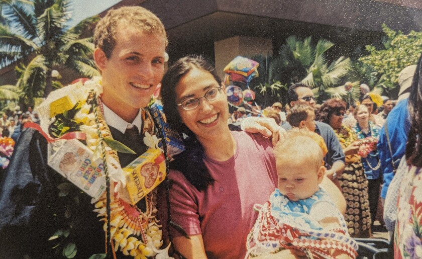 A woman with a baby stands next to a male graduate covered in leis.