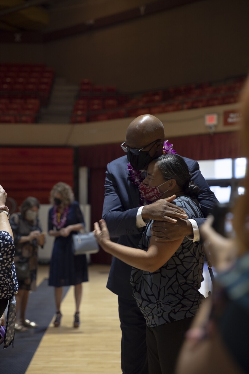 portrait photo of a tall man in a blue suit, purple lei and mask hugging a shorter woman with a red mask in the Cannon Activities Center