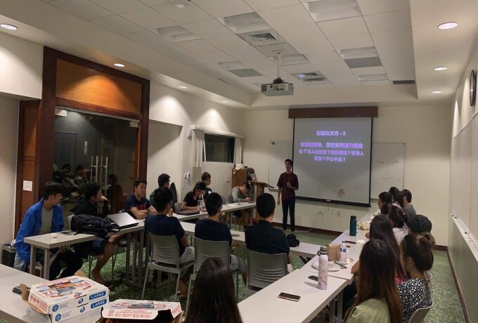 Students sit at tables and listen to a speaker in the front of the room