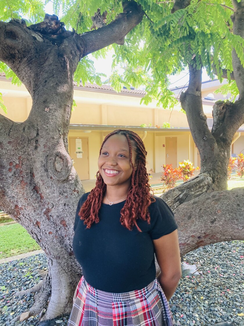 portrait of Black female student with red braided hair and a black shirt standing in front of the trunks of a tree in the BYUH art courtyard