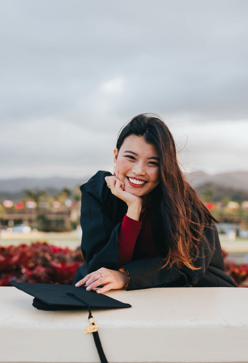 Natasya Haridas has one hand on her cheek and the other resting on the sign in the Flag Circle in front of BYUH. Her fingers are touching her graduation cap. She is smiling and wearing a red dress and black graduation gown.