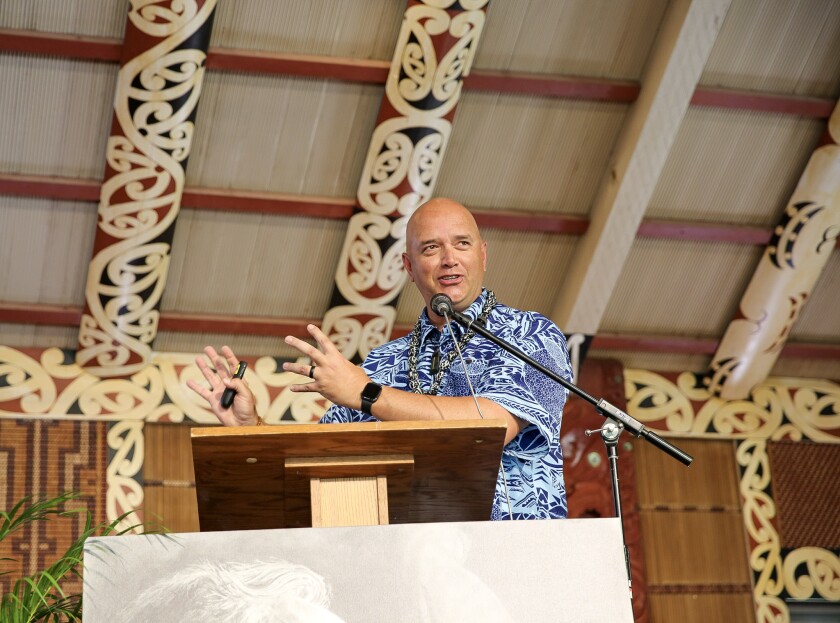 Wearing a blue and white aloha shirt and a kukui nut lei, BYU–Hawaii alumnus Seamus Fitzgerald, stands at a microphone in the Moari Marae or village at the Polynesian Cultural Center. The building has decorative painting, carvings and woven mats in it.