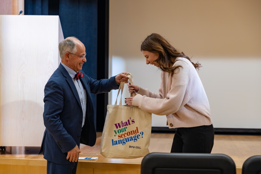 Dean Scott Miller hands MA student Kateryna Kravchenko a tote bag that says "What's your second language?" They are both smiling and standing in front of an auditorium stage.