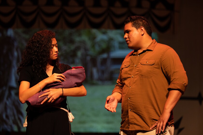Two actors on stage- a female wearing a black shirt holding a baby in a purple blanket, and a man wearing a mid-length orange shirt.