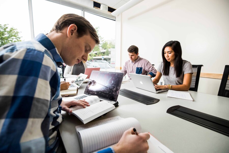 Students with laptops, books, notebook. Focus student is writing notes.