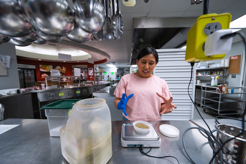 A woman in a pink shirt stands at a table in a metal kitchen. She looks down at a scale and measures a portion of spices.