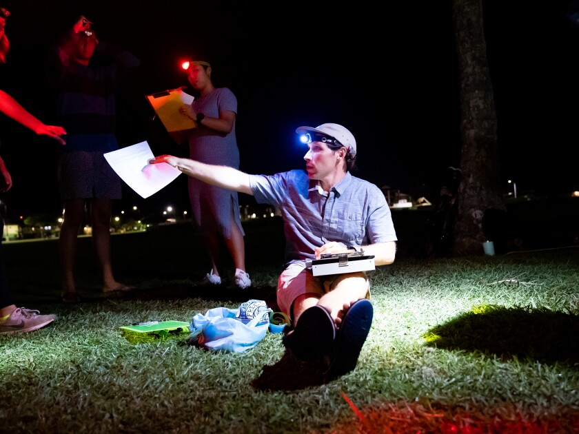 A man wearing a white hat and a head lamp with a blue short-sleeve button-up shirt and khaki shorts sits on the grass and is handing a paper to another student. It is completely dark except for where the man's headlight is shining.