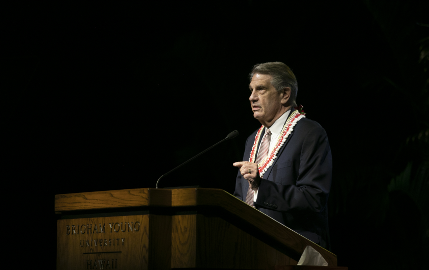 President Hallstrom wearing a dark suit and white and red poka dotted tie and white and red lei standing and speaking at a wooden podium with blackness behind him.