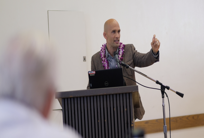 Mason Allred stands in front of a podium wearing a brown jacket and purple lei