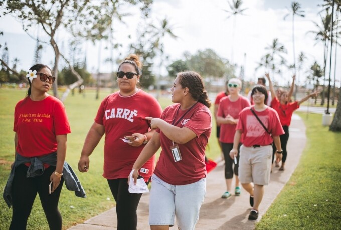 Students walk down the sidewalk wearing red shirts. There are palm trees in the background and grass on both sides of them.