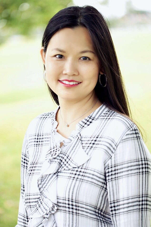 Dr. Hong smiles while wearing a plaid striped white and black blouse with greenery in the background.