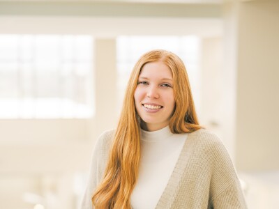 A pale woman with red hair wearing a beige shirt and cardigan smiles