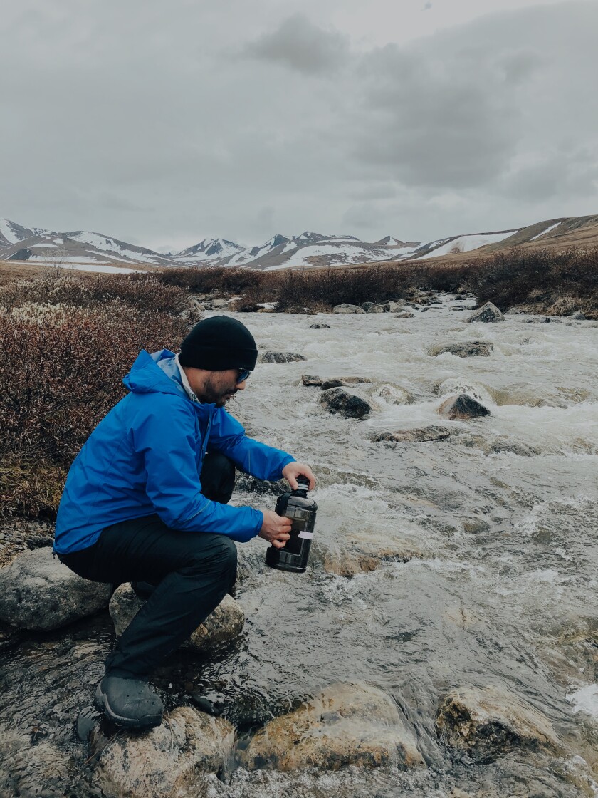 Raymond Lee is crouched on the banks of a running river. He holds a camera above the ground, capturing the permafrost. He's wearing a blue rain jacket, a black beanie, pants, and snow boots.