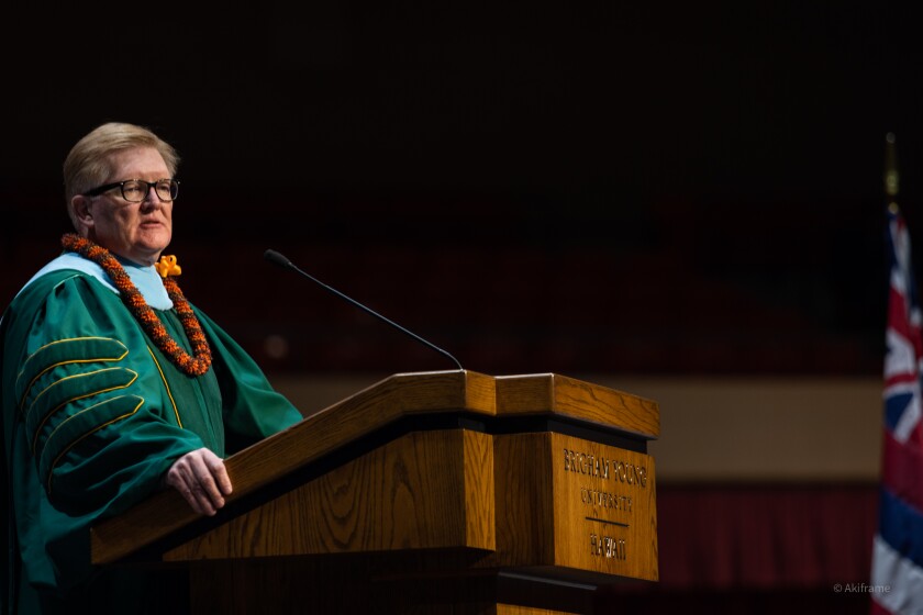 R. Kelly Haws, wearing an orange and brown lei with a blue, green and yellow gown, stands at the pulpit in the Cannon Activities Center.