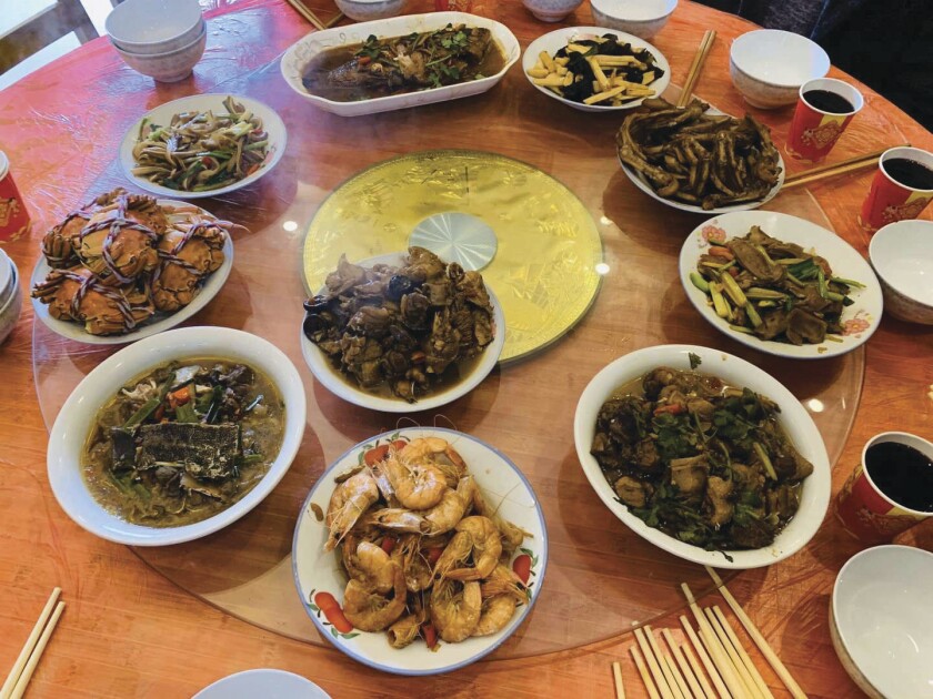 A large round table with a clear glass Lazy Susan holds 10 different Chinese dishes in white bowls and platters.
