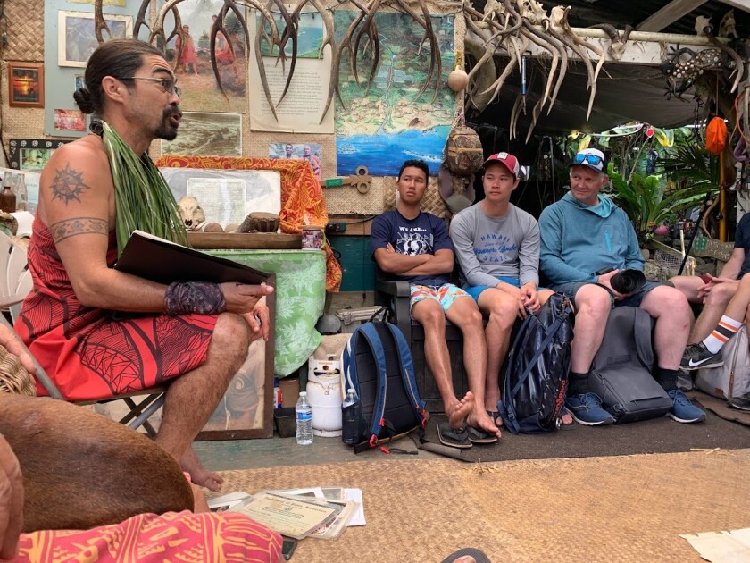 A man wearing traditional Hawaiian clothing sits on the left, talking to a group of teachers and students (Gideon Aiwohi, Kalai Ellis, and Richard Gill) on the right.
