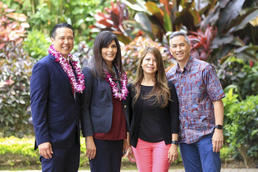 Taran K. Chun smiling next to his wife, both wearing suit jackets and pink flower leis, standing next to Vice President Jonathan Kau and his wife with greenery in the background.