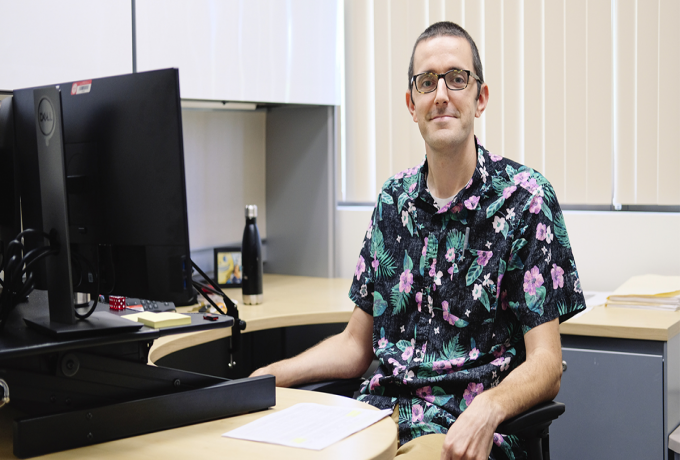 Man with glasses sits at a desk in an office.