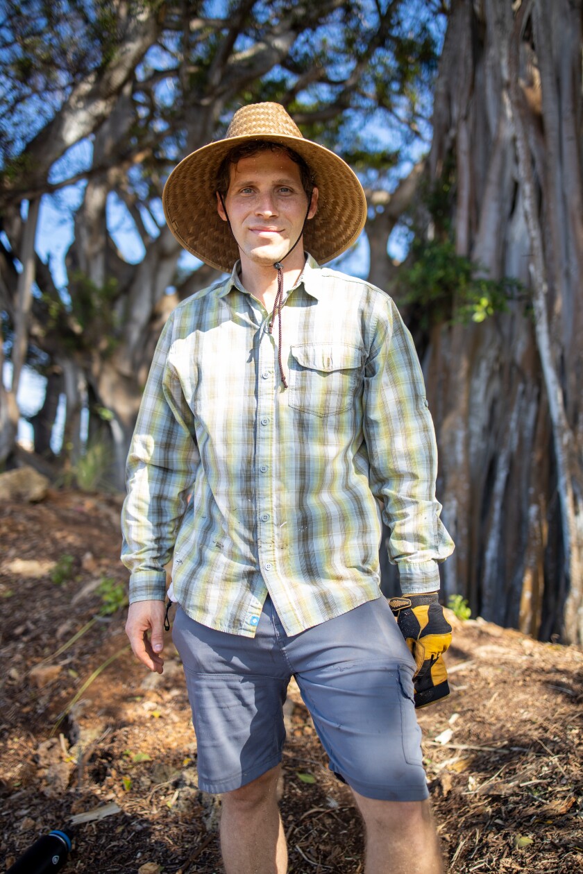Professor Ingley smiling wearing jean shorts, a plaid green and blue button-up shirt and a straw hat with trees behind him.