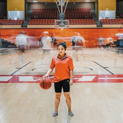 A girl is standing at the front of a gym wearing an orange shirt and black shorts. She is bouncing a basketball off the ground. There are people behind her moving around. Their bodies are blurred from the movement.