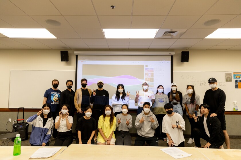 A large group of members of the Korean Club standing in a classroom in front of a white screen with a projected image of a song from YouTube on it. They are all wearing masks.