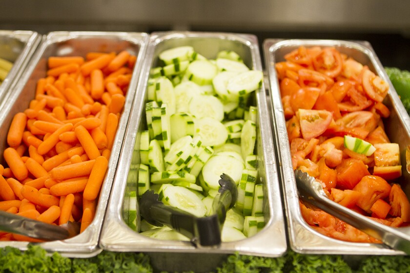 landscape close-up photo of three silver food hotel pans of orange carrots, green cucumber slices and red tomatoes with silver and black tongs in each pan at the cafeteria