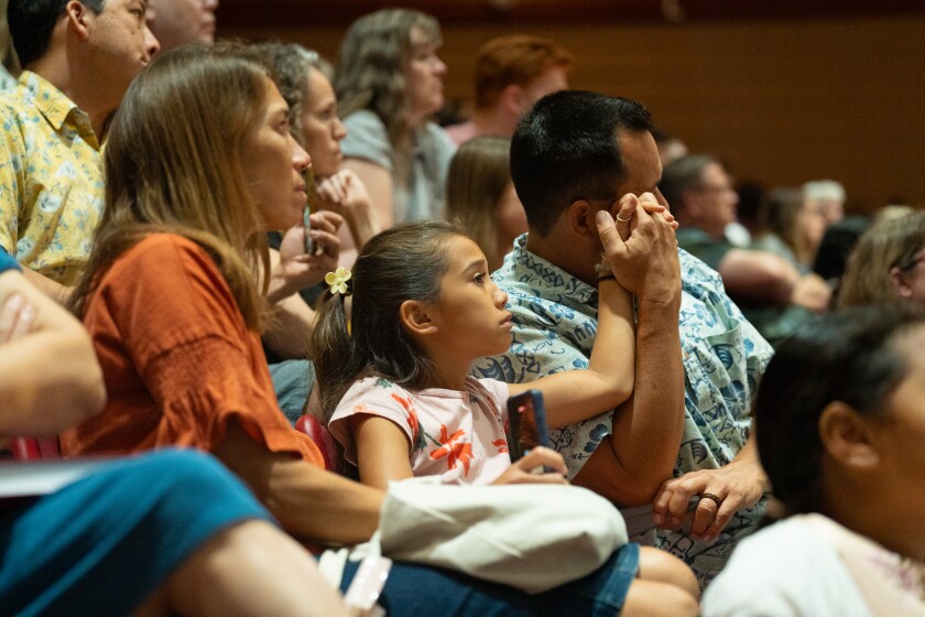 A mother, daughter, and father sitting in a crowd watching a performance. The father holds the daughters hand up to his cheek as they both watch.