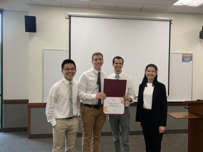 three men in white suits with ties and one business formal dressed woman smiling at a camera and holding up a certificate in a classroom