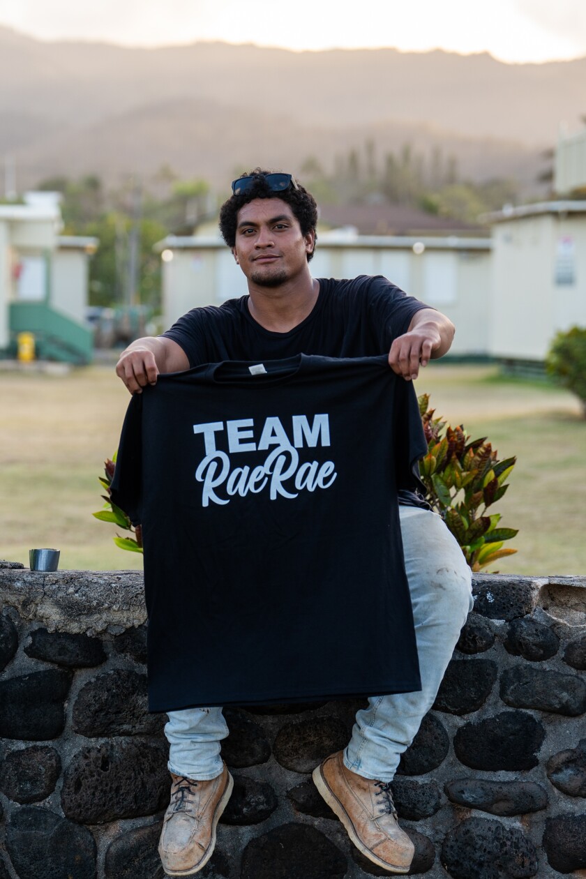 A man sitting on a stone wall holds up a t-shirt that says "Team Rae Rae"