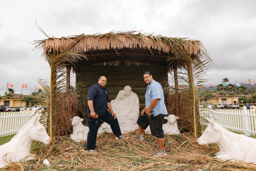 Two contractors, one wearing navy blue polo and pants, and the other wearing black shorts and a light blue short-sleeve button-up shirt, stand in front of the BYUH nativity facing the camera.