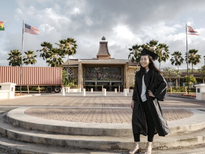 Muugii Galbadrakh smiling standing wearing a black graduation gown and cap with the BYUH campus and flags behind her.