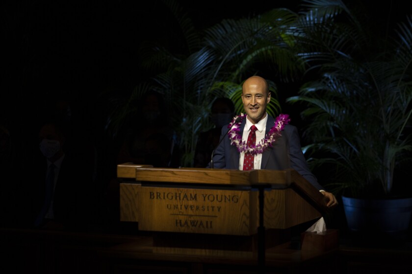 landscape shot of man in suit and red tie wearing a purple lei standing at the pulpit in the Cannon Activities Center giving his devotional address