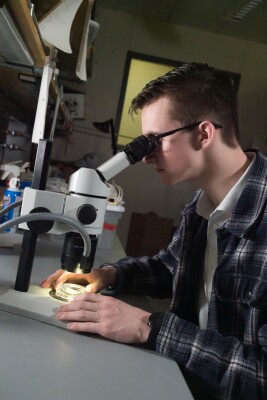 A pale man with brown hair and glasses wearing a plaid shirt looks into a microscope.