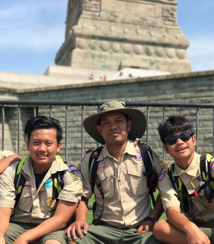 Saborn Va sitting in the middle of two young male refugee scouts. All three in scouting uniform in front of a statue in New York City.