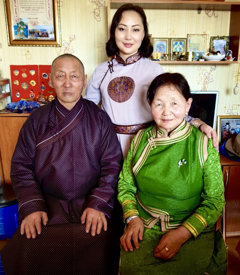 Bazarragchaa standing next to her parents sitting down on chairs in traditional Mongolian clothing in their house.