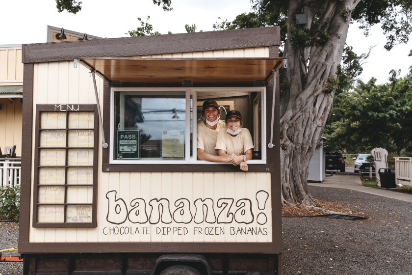 A couple wearing baseball caps and beige T-shirts stands in the window of their frozen banana food truck in Haleiwa.