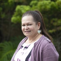 A side portrait of Greta Damuni smiling at the camera with blurred green plants in the background. She has brunette hair in a ponytail, wearing white dangly earrings and purple outerwear.