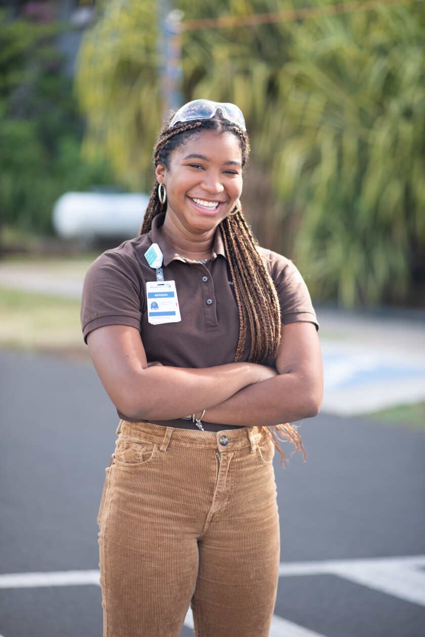 Mathurent smiling wearing seethrough medical glasses, a name tag on her brown shirt and brown pants and folding her arms with greenery and pavement behind her.