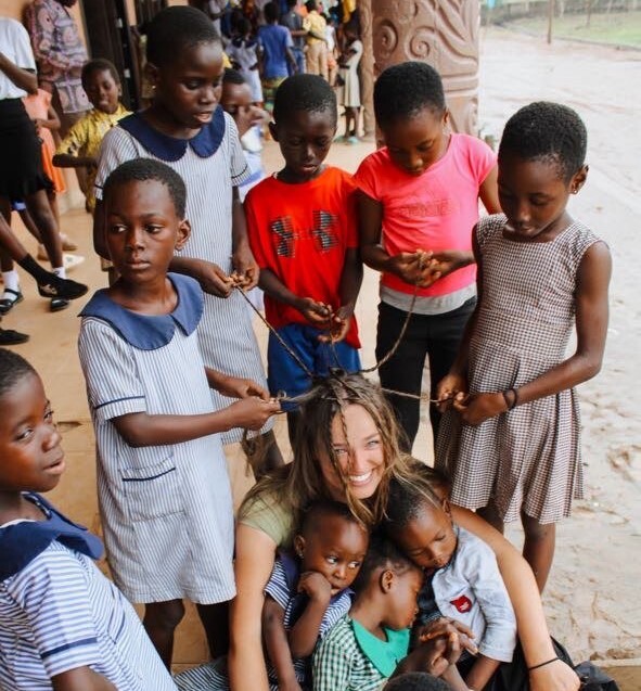 A girl with light brown hair is getting her hair braided by a group of African kids. Other kids are asleep in her lap.