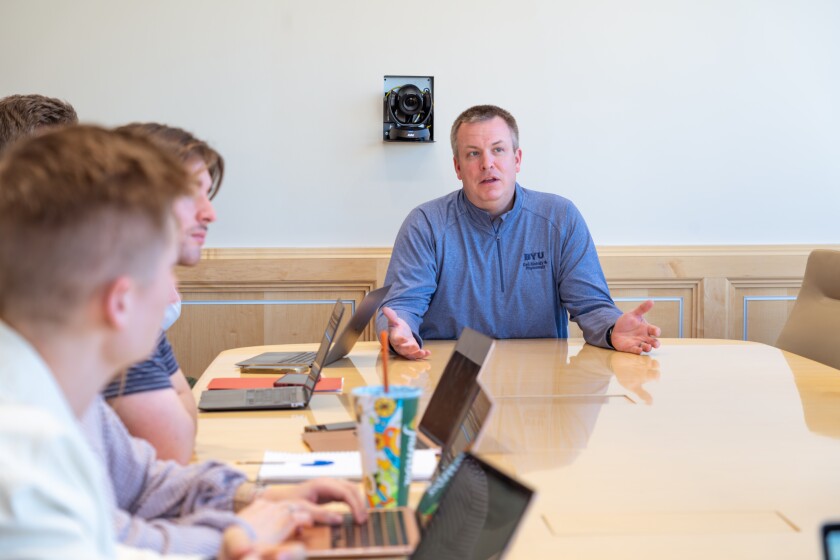 A male professor wearing a blue BYU pullover sits at the front of a table talking with students