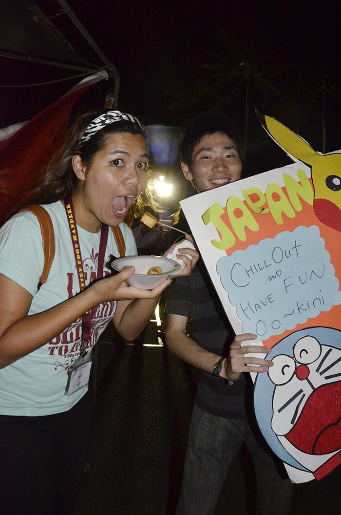 vertical image of two students together at night, one female with a styrofoam bowl putting food into her mouth and the other, male, holding a poster that says "JAPAN chill out and have fun" with various drawings and colors on it