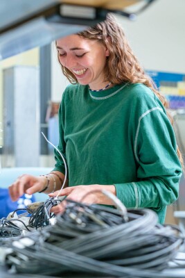 A woman grins as she picks up a bundle of wires, larger piles in the foreground partially blocking her body. She has light skin and chest-length curly brown hair falling down her back. She wears a long-sleeve plain green shirt, the sleeves pushed back to her elbows, several beaded bracelets on her right arm in the background, and a blue and white beaded necklace.