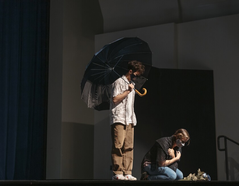 On a black stage, there is a man wearing tan pants, a white button-up shirt that's not buttoned with a white shirt underneath holding a black umbrella and a woman wearing a black shawl kneeling over on the ground with flowers in front of her.
