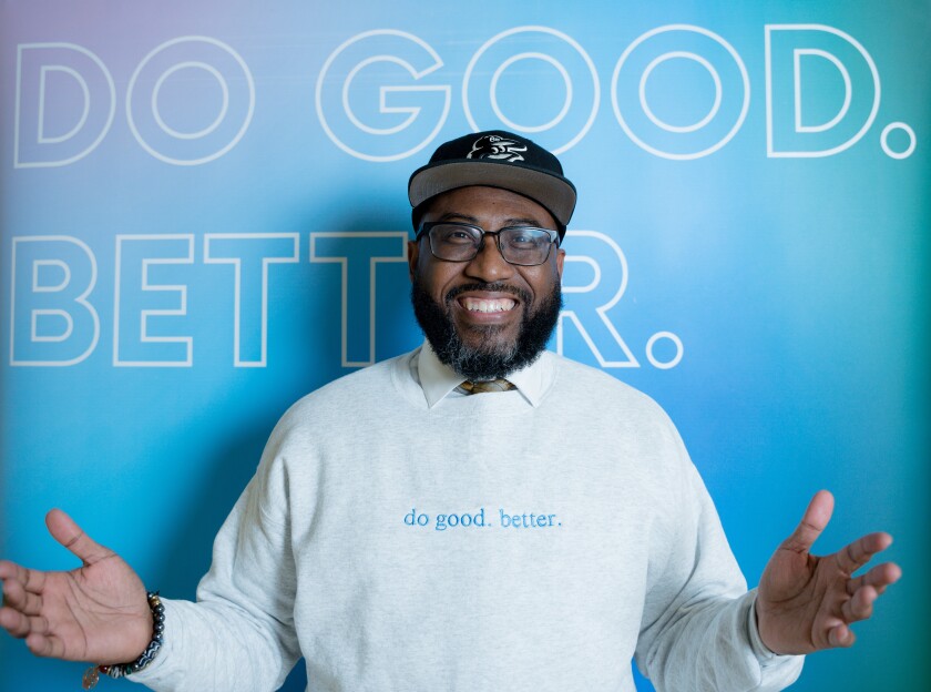 Reverend Dr. Heber Brown III smiles in front of a blue "Do Good. Better." sign wearing a gray sweatshirt with blue embroidered letters that read "do good. better."