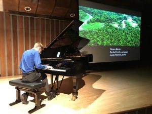 A young man in a blue button-up shirt plays piano with an image of the Amazon forest behind him.
