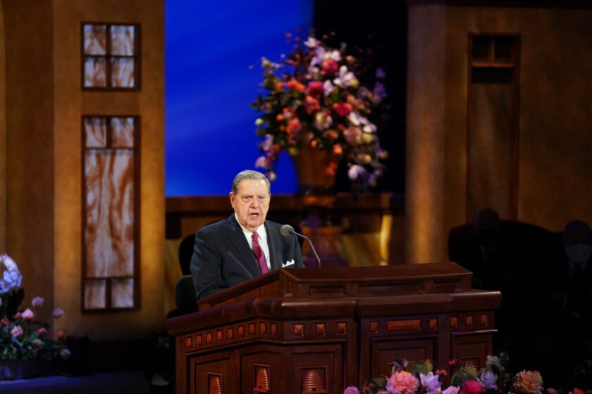 Elder Jeffrey R. Holland  speaking at a wooden pulpit with tan and brown walls with a big blue window and flowers behind him.