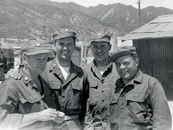 Nelson and friends stand in military uniforms in a black and white photo with mountains in the background.